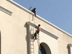 Painter dangling by two ropes on a wooden ladder while another worker lowers him a can of paint. They work with no net, no harness, no nothing at this third story height.